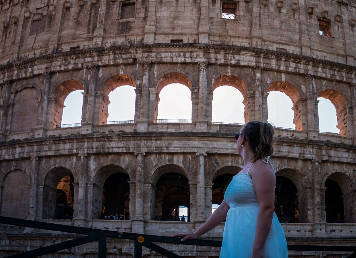 A woman in a white dress gazes at the sunlit arches of the Colosseum, conveying wonder and awe. The ancient structure towers majestically in the background.